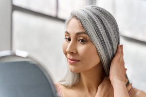 Senior happy middle aged mature asian woman closeup portrait. Skin care advertising. Portrait of beautyful happy middle aged mature asian woman, senior older 50s lady pampering touching gray hair looking at herself at mirror indoors. Ads of lifting anti wrinkle skin hair care spa.