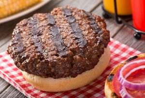 A juicy barbecued hamburger with grill marks on a rustic picnic table with tomato, onion, mustard, ketchup, and corn.