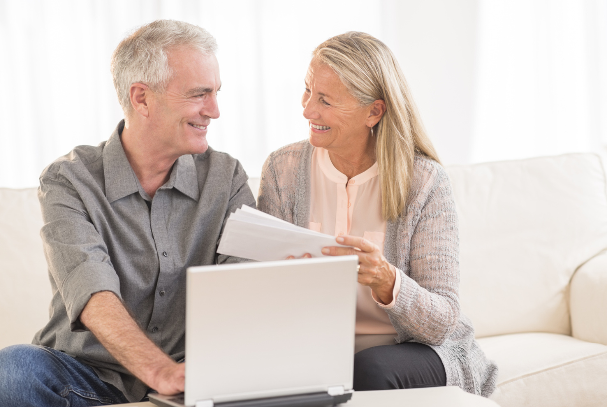 Boomer couple turning 65, smiling at each other in fron of an opened laptop.