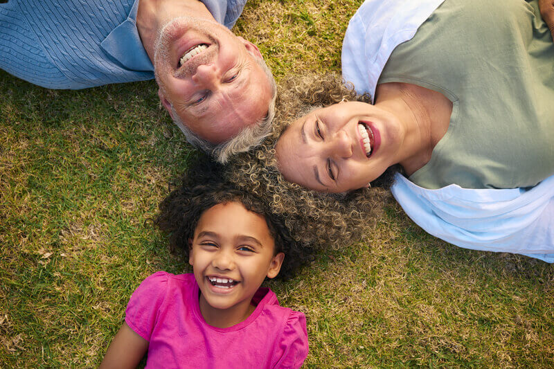 Looking Down On Multi Racial Grandparents With Granddaughter Lying Outdoors On Grass