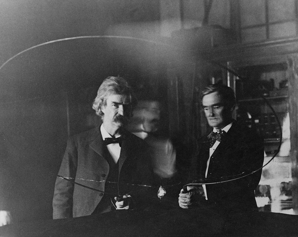 Serbian-American inventor and engineer Nikola Tesla (1856 - 1943, blurred at centre) performs an electrical experiment for writer Samuel Langhorne Clemens, aka Mark Twain (1835 - 1910, left) and actor Joseph Jefferson (1829 - 1905), 1894. They are at Tesla's laboratory at 35 South Fifth Avenue, New York City, and an electrical current is being passed through Twain and Jefferson, in order to illuminate a lamp.