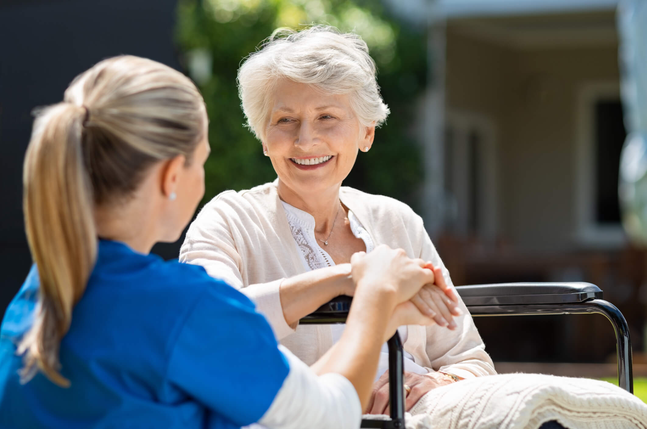 Nurse takes care of old patient. Smiling senior patient sitting on wheelchair with nurse supporting her. Doctor looking at elderly patient on a wheelchair in the garden. Nurse holding hand of mature women outside pension home.