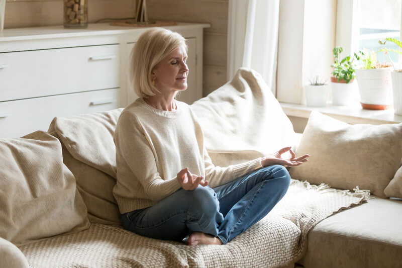 Mindful healthy mature woman practicing yoga meditation at home. Mindful healthy mature woman practicing meditation at home, calm senior middle aged lady sitting on couch in lotus pose doing yoga for mental balance breathing air relaxing on stress free weekend