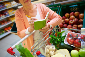Shopper with notepad. Image of senior woman looking at product list with goods in cart near by. Medicare grocery card.
