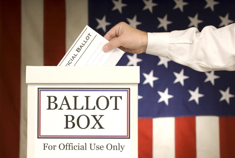 Ballot Box With Hand Voting. Male hand putting ballot in the ballot box. American Flag background.