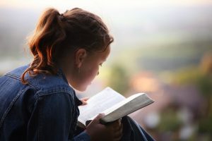 Cute girl is reading the bible in the evening sun on a small hill over the country.