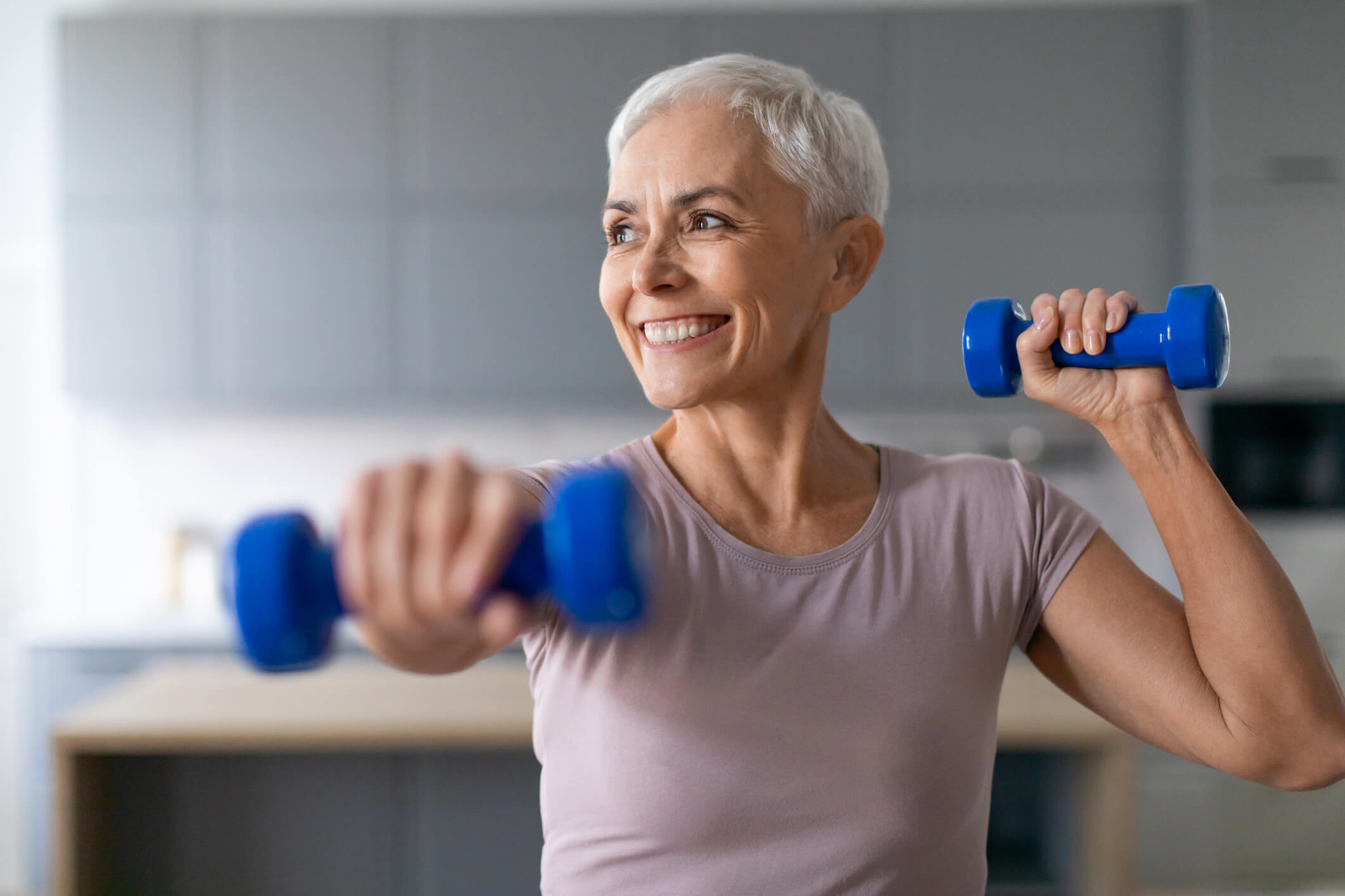 Senior woman enjoys morning workout with weights in living room. Smiling senior woman enjoys morning workout with weights, lifting dumbbells exemplifying health and wellness in her living room at home. Sporty lady exercising during domestic training