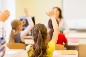 Group of school kids raising hands in classroom. Education, elementary school, learning and people concept - group of school kids with teacher sitting in classroom and raising hands