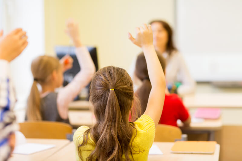 Group of school kids raising hands in classroom. Education, elementary school, learning and people concept - group of school kids with teacher sitting in classroom and raising hands