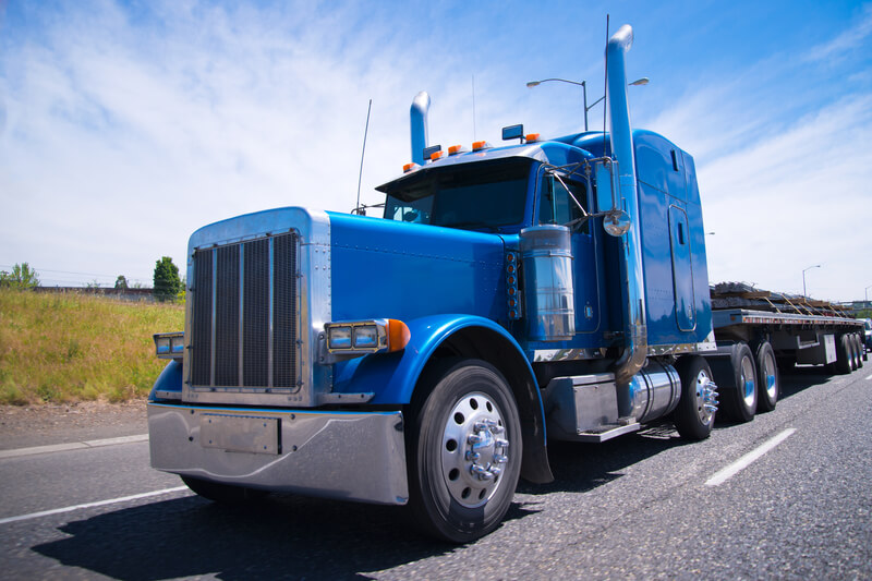 Big rig semi truck blue wolf of roads. Classic blue bonneted big rig semi truck with chrome accessories and details on a multilane highway road with a laden flat bed trailer on background of blue sky.