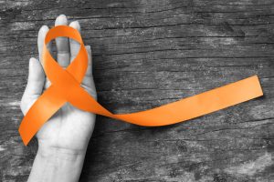 Orange ribbon for Leukemia, Kidney cancer, RDS multiple sclerosis awareness on human hand, aged background; Satin fabric color symbolic concept for raising public support on people living with disease.