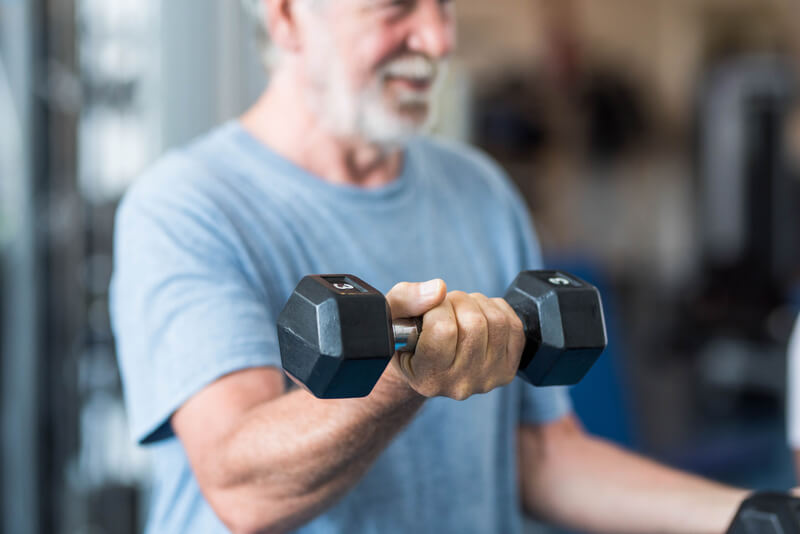 Close up of mature man holding two dumbbells doing exercise at the gym to be healthy and fitness - portrait of active senior lifting weight.