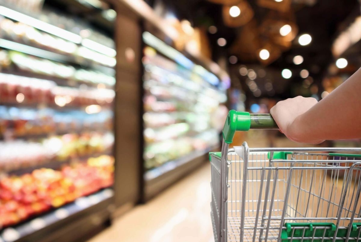 Woman hand hold supermarket shopping cart with Abstract grocery store blurred defocused background. Woman hand hold supermarket shopping cart with abstract blur organic fresh fruits and vegetable on shelves in grocery store defocused bokeh light background