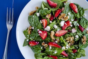 Fresh organic strawberry spinach salad with walnuts and feta cheese and poppy seed dressing on white plate sitting dark blue background and fork on side