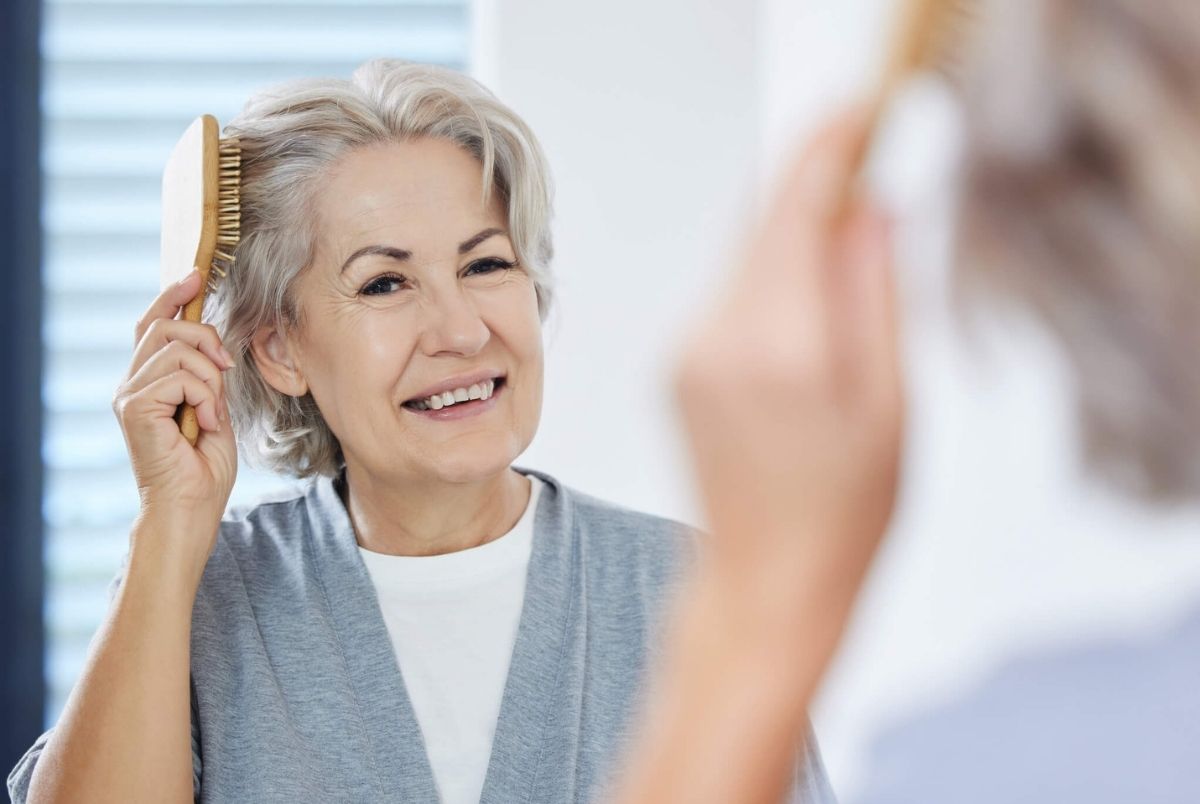 Home, senior woman and comb hair in mirror for maintenance, texture and growth in bathroom. House, reflection and elderly person with hairbrush for scalp health, wellness and self care in retirement.