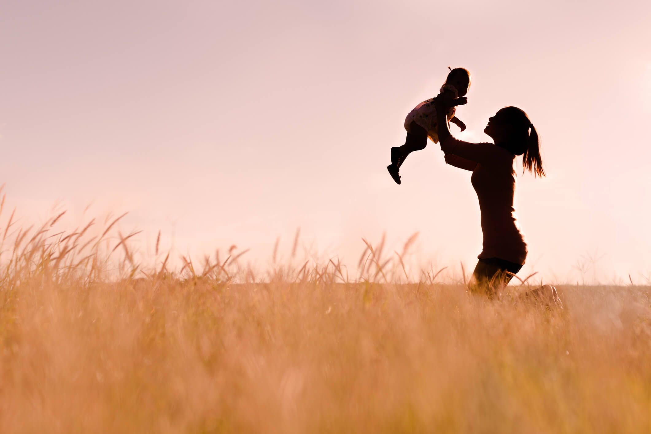 Mother playing with her baby outdoor, silhouette. A mom lifting her child in the air having fun sitting on the soft grass outside in the field