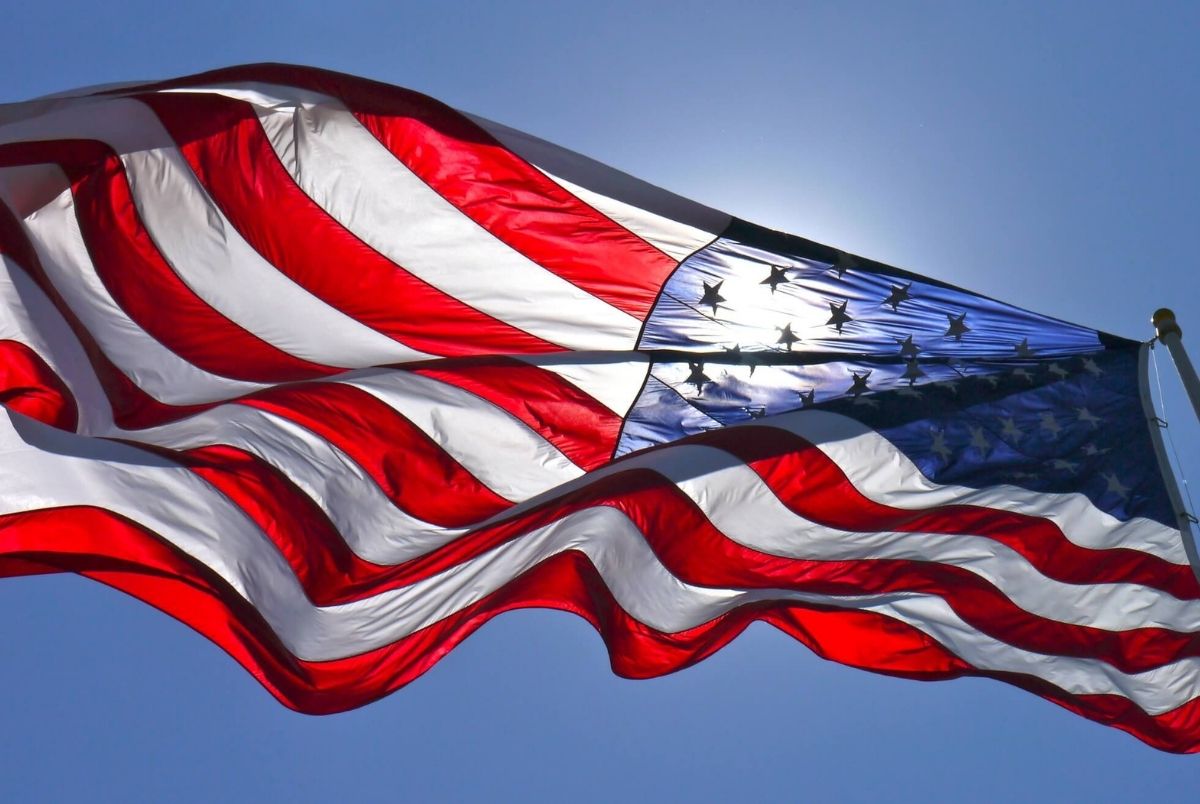 Sun shining through American flag flying in blue sky.