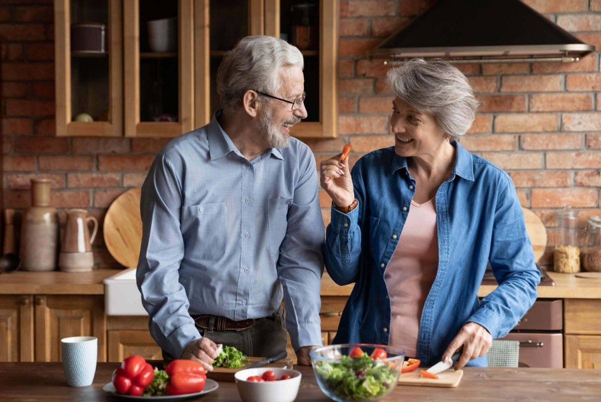 Older couple cooking together healthy food in modern kitchen. Older couple cooking together vegetarian food in kitchen. Mature spouses look at each other with love prepare vegetable paprika natural dish. Food delivery order clients, healthy eat lifestyle concept