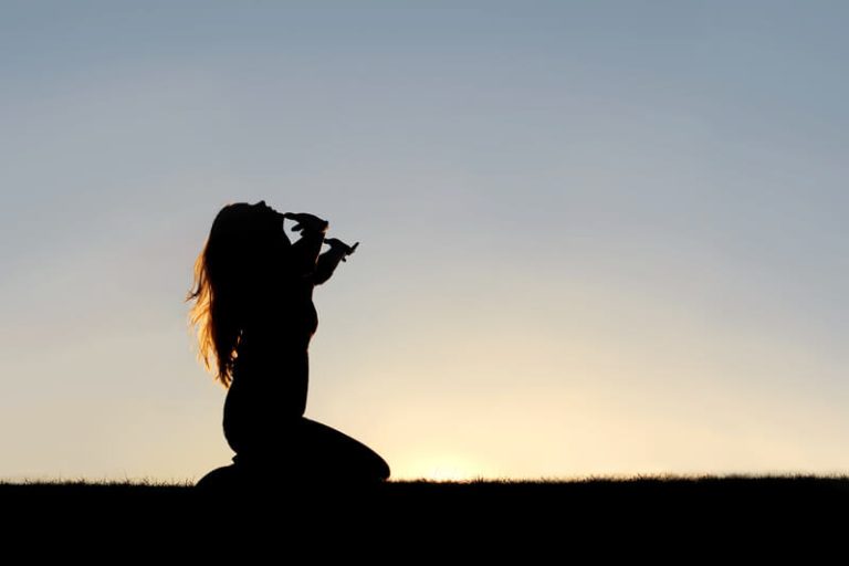 Silhouette of Woman Kneeling in Prayer and Surrender. A silhouette of a woman kneeling down with her hands in the air, praying, thanking, and surrendering to God.