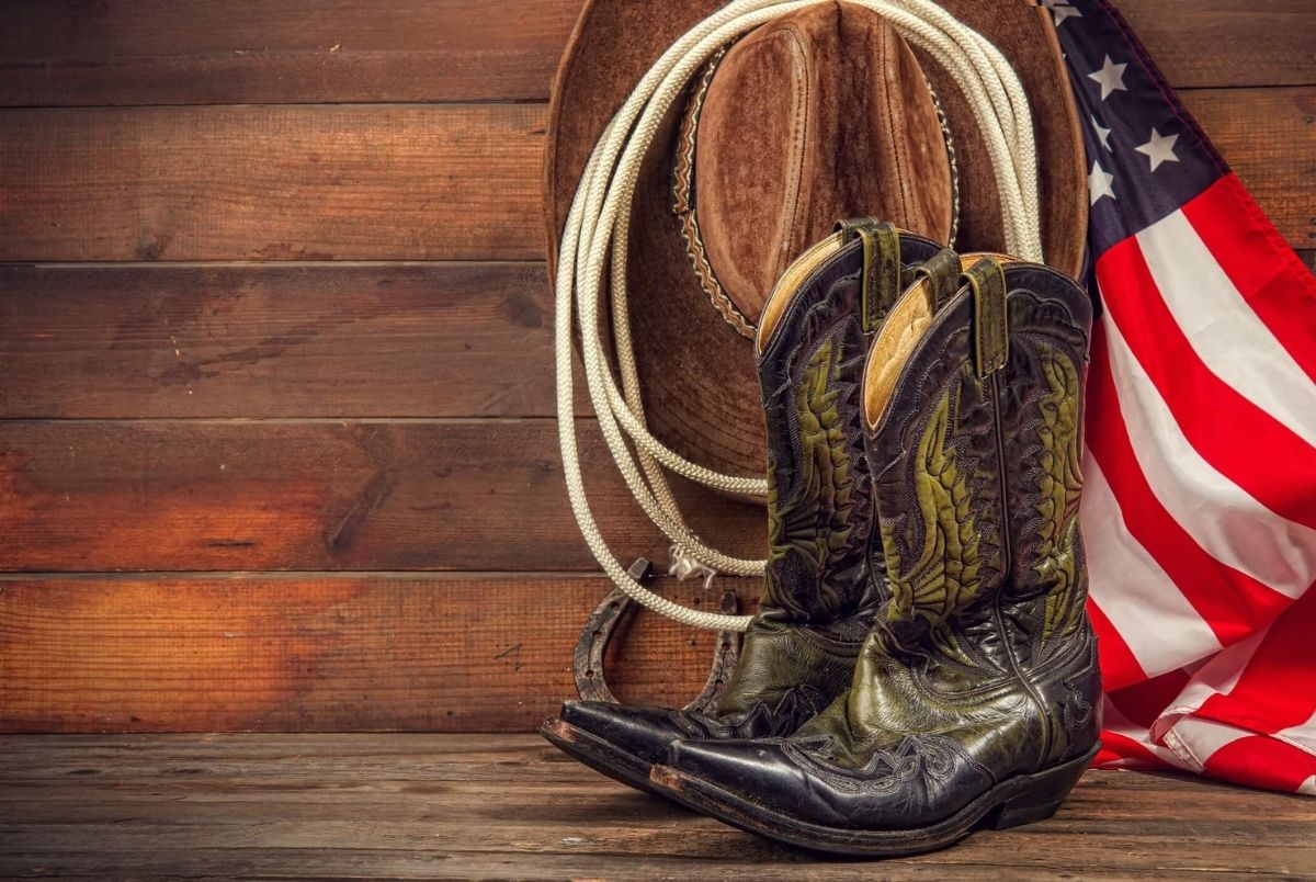 Wild west still life. Shabby old ornate classic cowboy boots hat and lasso on wooden background and USA flag background