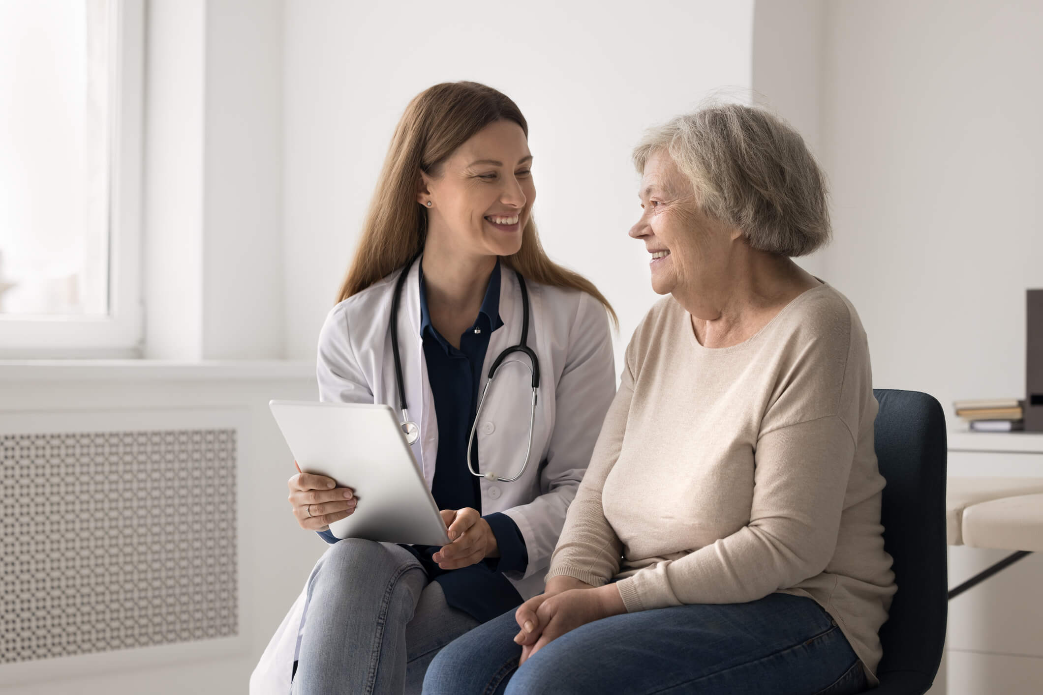 Smiling young doctor examine elderly patient make records on tablet. Your results are good. Smiling friendly young female doctor attending physician examine elderly women make records to patient medical file on tablet pc see positive dynamics compared to previous tests