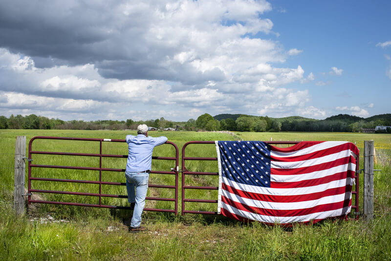 Man standing at metal gate in beautiful grass field, American flag hanging on fence, blue sky white clouds