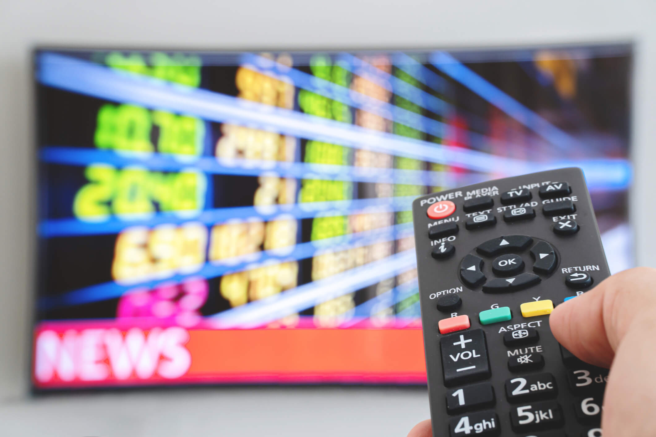 Hand holding TV remote control with a television news in the background. The man switches the news channels on the remote control on TV