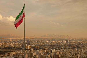 Iran Flag in the Wind Above Skyline of Tehran. Iran flag waving in the wind above skyline of Tehran lit by orange glow of sunset.