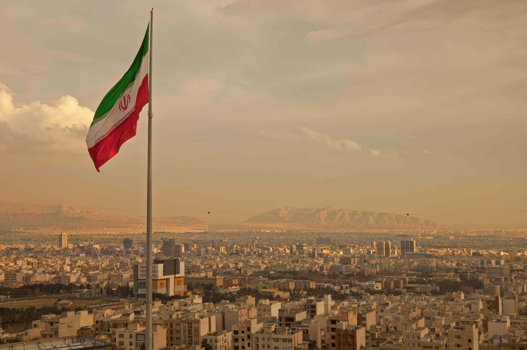 Iran Flag in the Wind Above Skyline of Tehran. Iran flag waving in the wind above skyline of Tehran lit by orange glow of sunset.