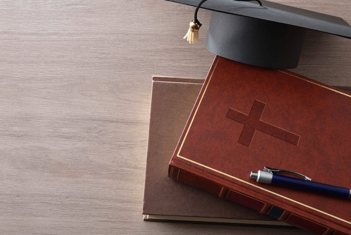 Religious education with Bible books and mortarboard on wooden desk. Religious and educational background with a Bible on a textbook, pen, and cap on a wooden desk. Top view