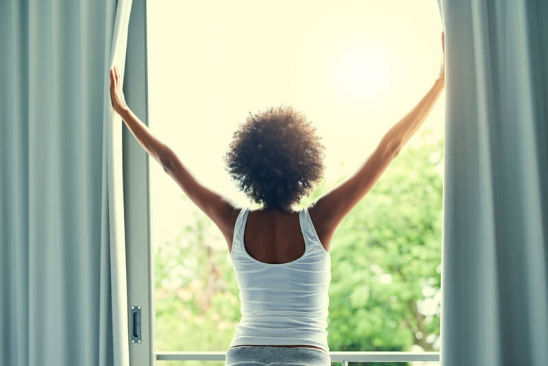 Back, window and curtains of girl for wake up, bright light and wellness by bedroom in morning. Home, female person and fresh air after sleeping, resting and sunshine on vacation in California.