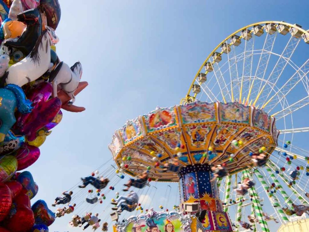 A Wellenflug (wave swinger) ride spinning patrons high above a carnival ground with a massive ferris wheel rotating in the background.