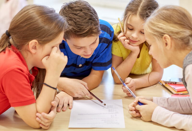 Group of students talking and writing at school. Education, elementary school, learning and people concept - group of school kids with pens and papers writing in classroom